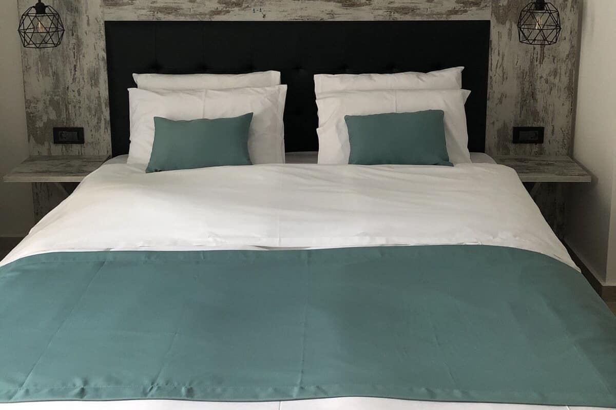 A neatly made bed with white and teal bedding, a dark headboard, and two bedside tables with geometric wall-mounted lamps.