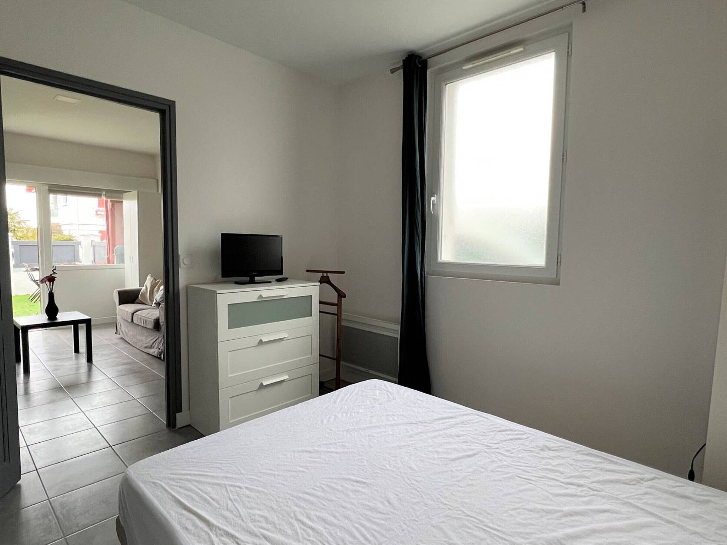 Een slaapkamer met een bed, een televisie op een dressoir, een raam en een deur die leidt naar een andere kamer.