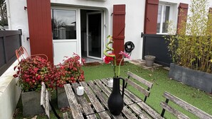 Un patio con una mesa de madera, un jarrón negro con flores y un banco. Hay una puerta con el cartel “ARTATEGIA” encima.