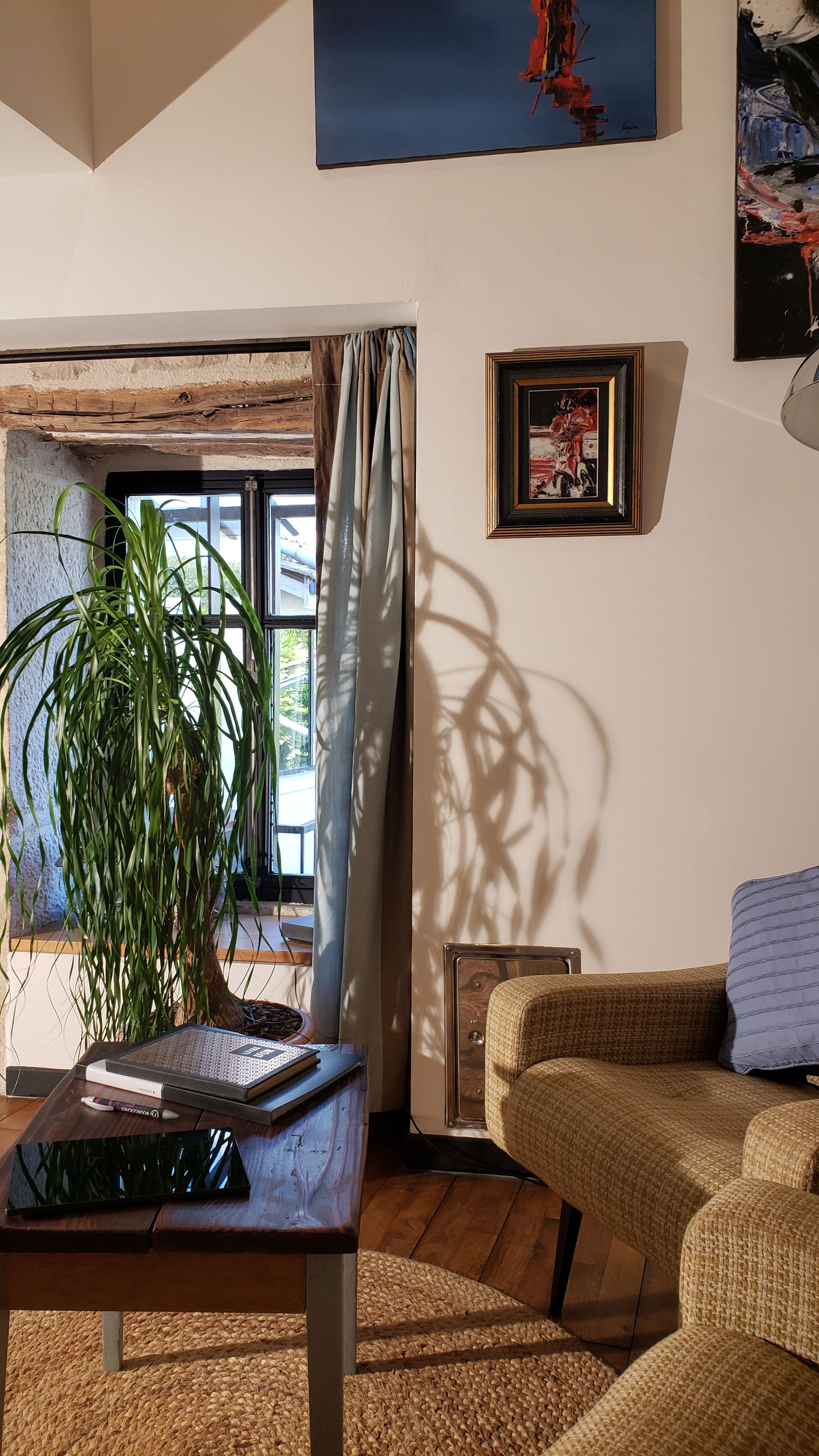 A living room with a sofa, a table with a book and a pen, a potted plant, a window with curtains, and a painting on the wall.