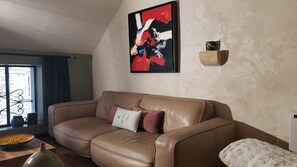 A living room with a brown leather sofa, a colorful abstract painting, a stone accent piece, and a view through a window. - L'autre Bout de Toit (Meursault)