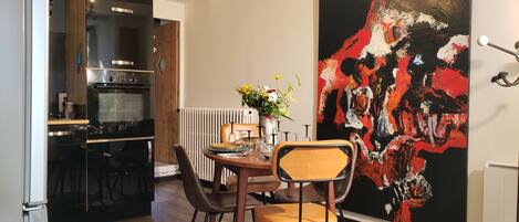 Une cuisine moderne équipée d’un four Bosch, un coin repas avec une table dressée pour quatre personnes, et un grand tableau abstrait accroché au mur.