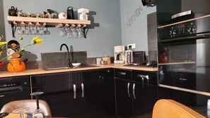 A modern kitchen with black cabinets, a Bosch oven, a coffee maker, and a shelf with jars and glasses. - L'autre Bout de Toit (Meursault)