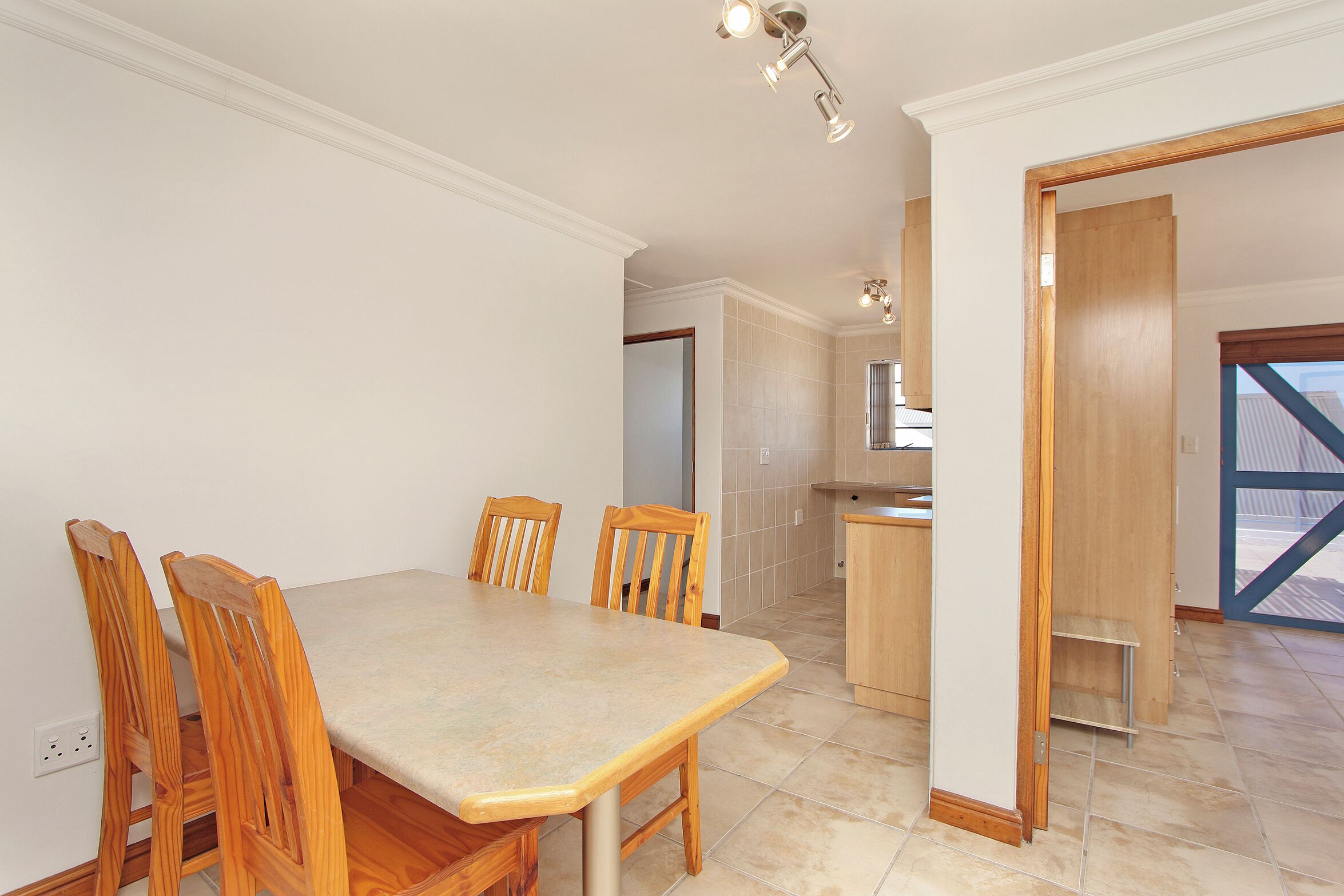 A dining area with a table and chairs, a kitchen in the background, and a door leading to another room.