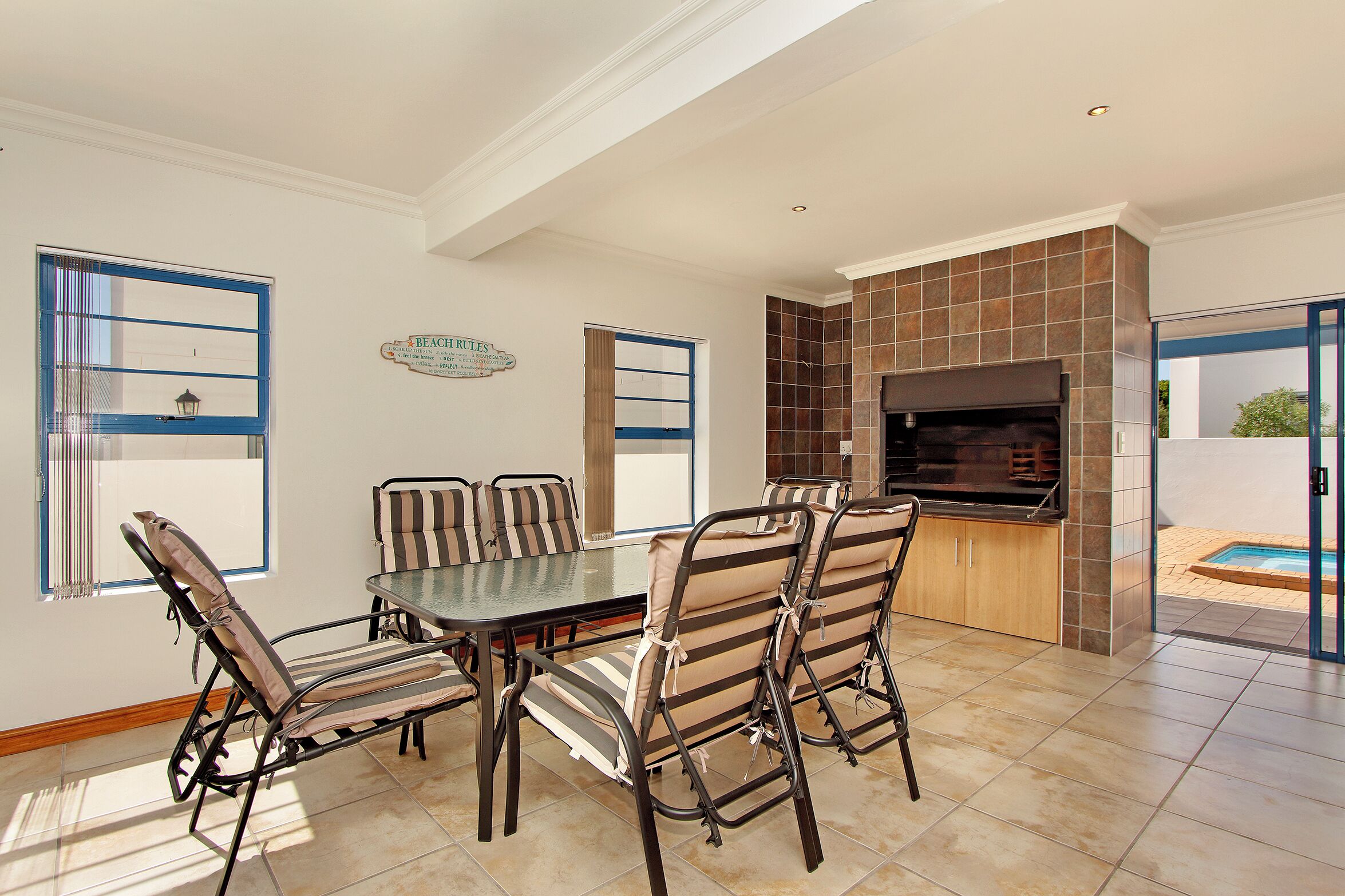 A dining area with a glass-top table and striped chairs, a built-in grill, and a view of a swimming pool.