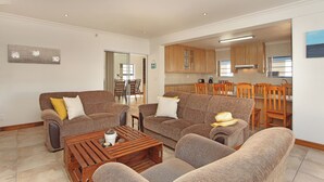 Een ruime woonkamer met twee banken, een houten salontafel, en een eethoek met houten stoelen. Op de achtergrond is een keuken te zien.