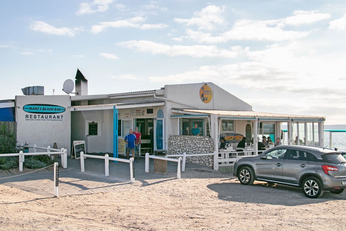 Un restaurant situé en bord de mer, avec une enseigne, un espace extérieur abrité avec des tables et des chaises, et une voiture garée devant.