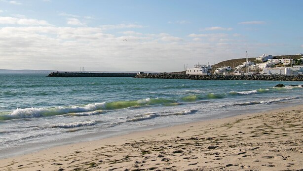 Een kuststrand met een rotsenrij, kalme zee en witte gebouwen met een glooiend achtergrondlandschap.