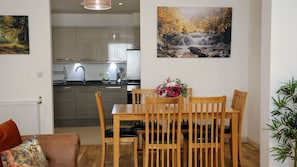 A dining area with a wooden table and chairs, a vase of flowers, and a kitchen in the background.