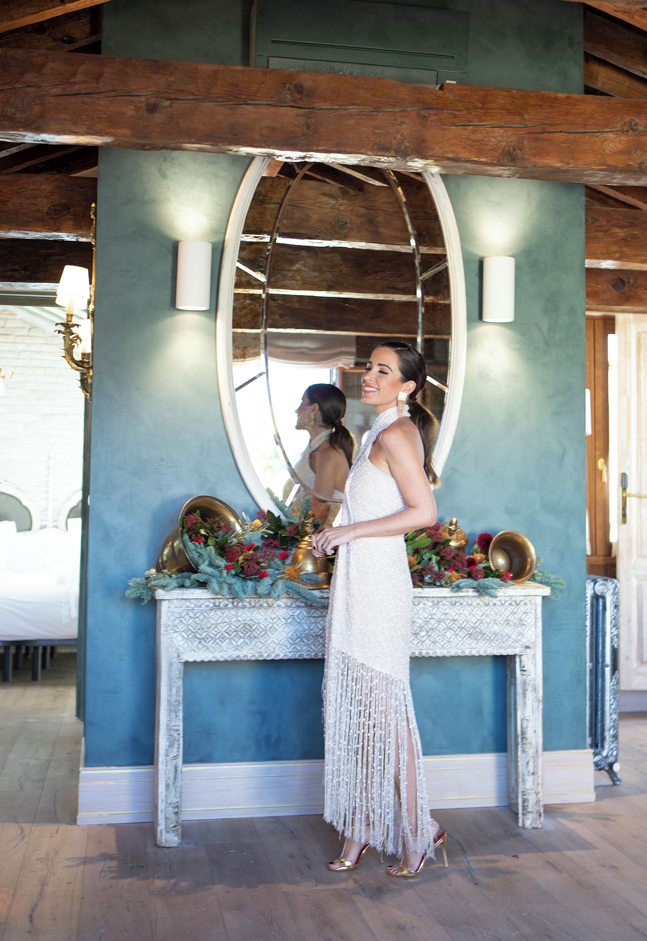 Una mujer con vestido blanco de pie frente a un espejo con una consola decorativa.