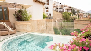 Una piscina en la azotea con borde de azulejos, rodeada por un muro de piedra bajo, mobiliario de mimbre y plantas en macetas.