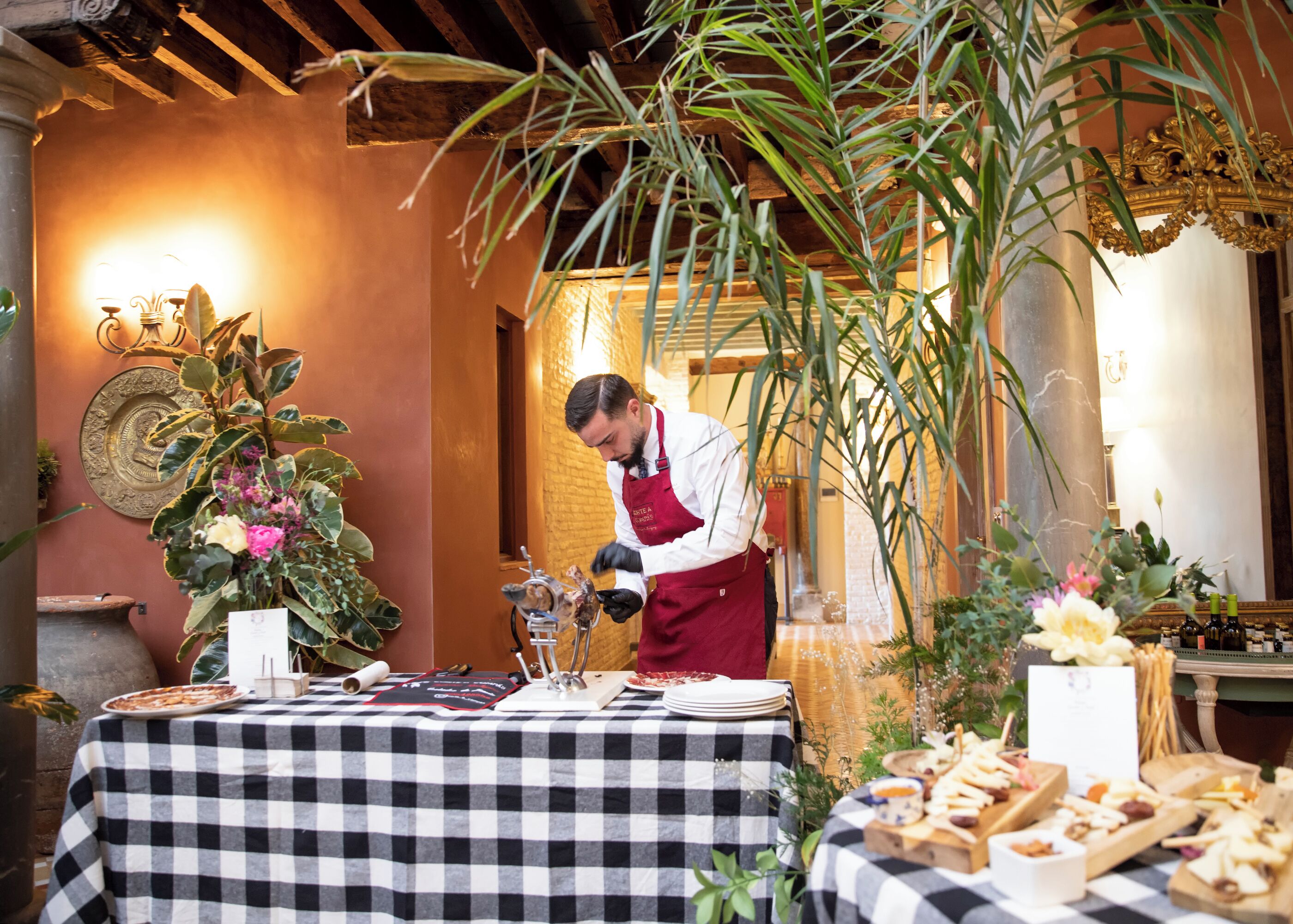 A chef preparing food at a table with a checkered tablecloth, surrounded by plants and a decorative wall.