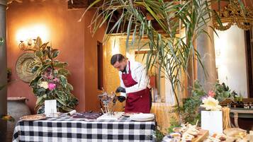 A chef preparing food at a table with a checkered tablecloth, surrounded by plants and a decorative wall.