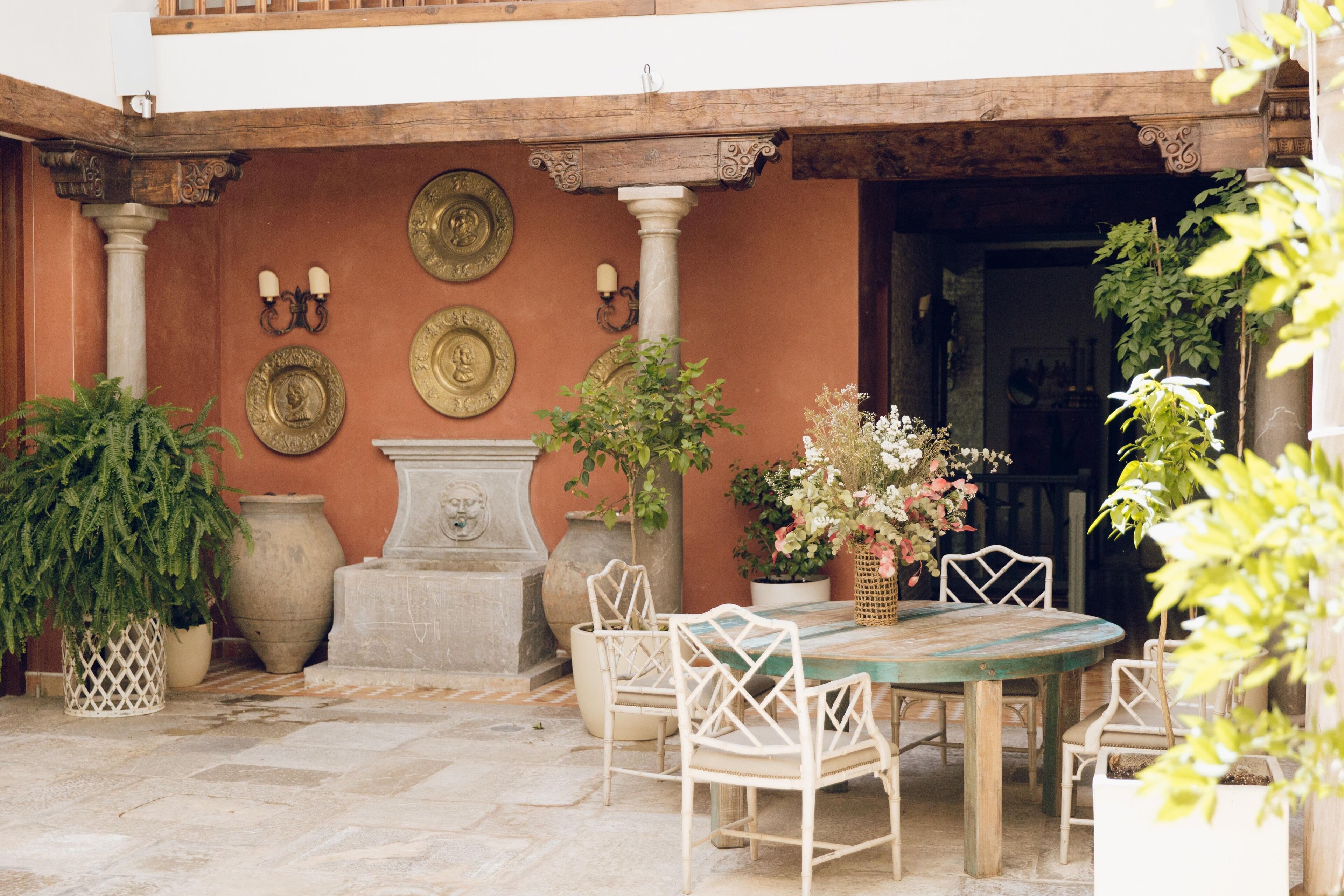An outdoor patio with a round table and white chairs, surrounded by potted plants and decorative plates on the wall.