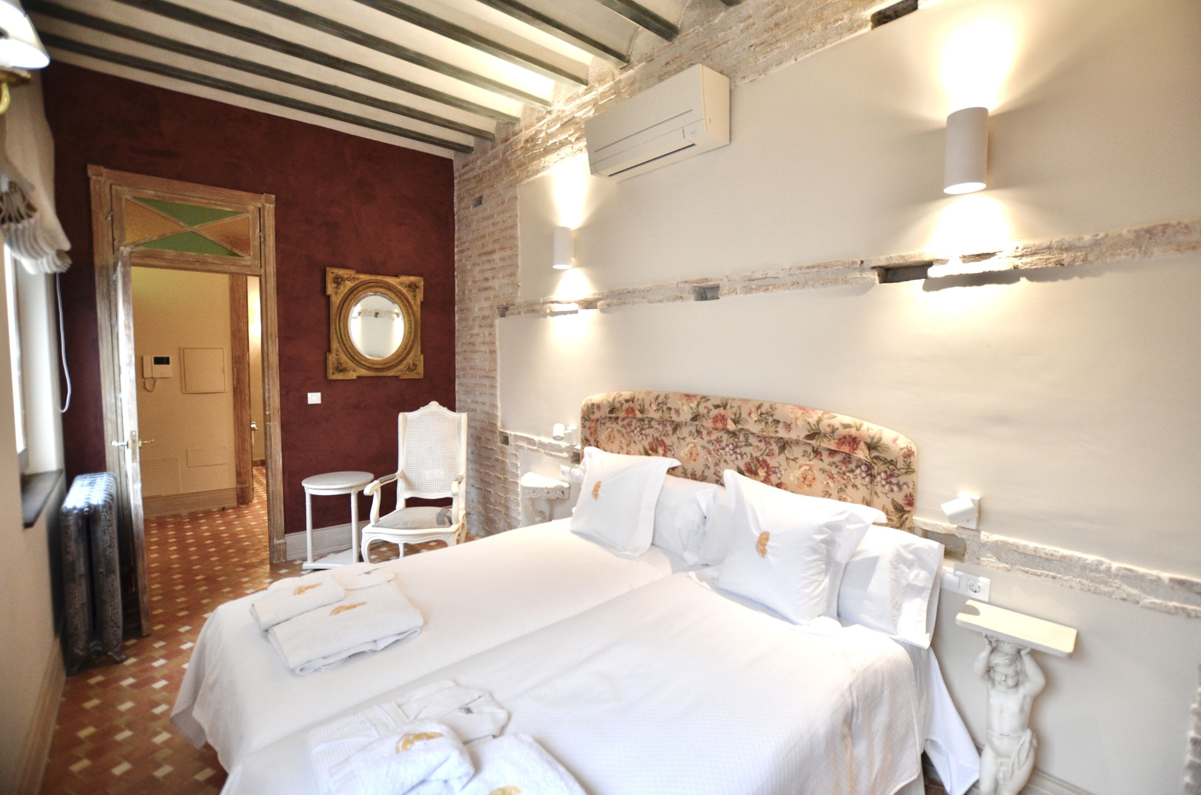 Une chambre d’hôtel avec un grand lit, une tête de lit à motifs floraux, un miroir et un mur blanc agrémenté d’un décor mouluré.