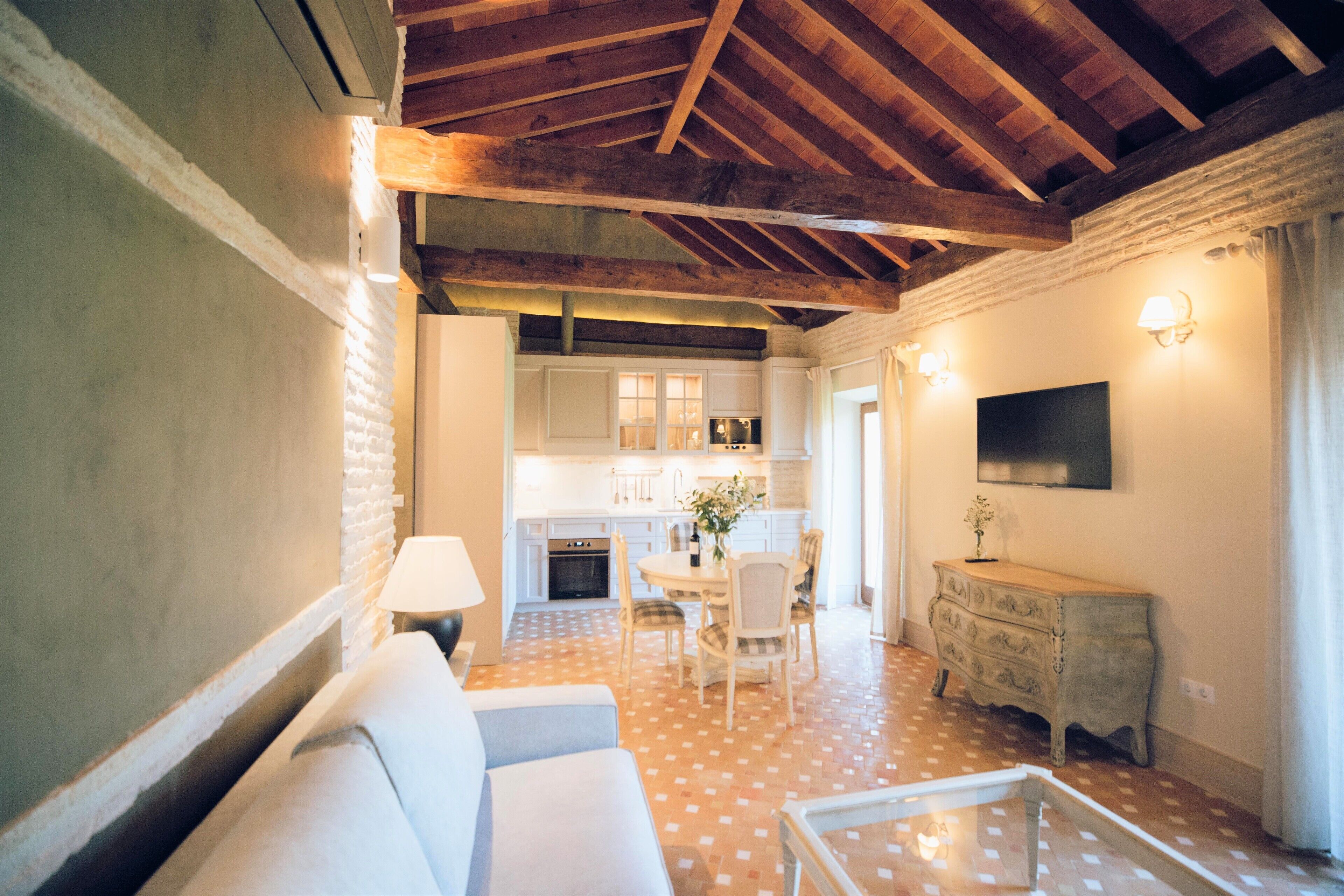 A cozy living room with a sofa, a dining table with chairs, a kitchen in the background, and a TV mounted on the wall.