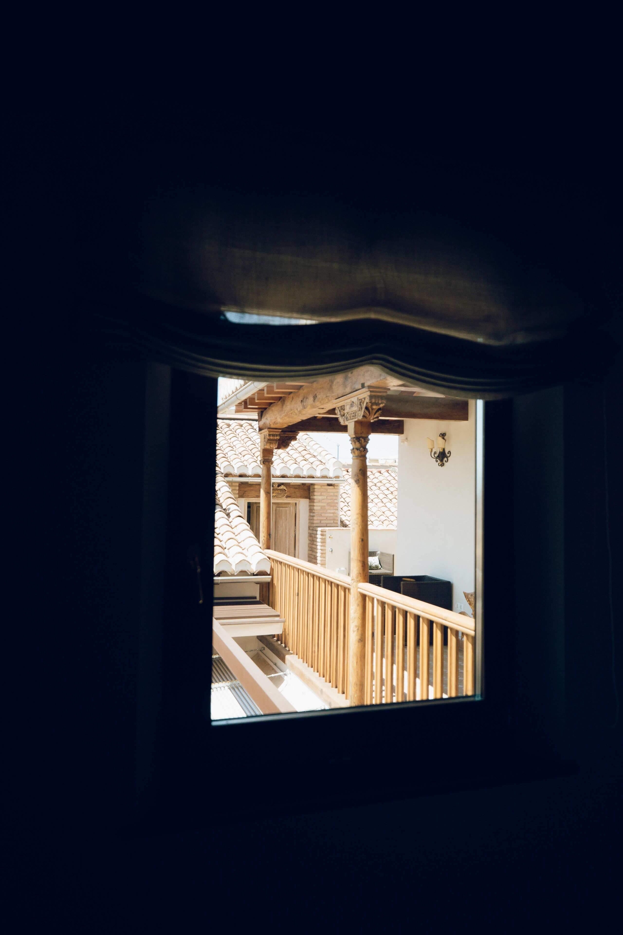 Vista a través de una ventana que muestra un balcón de madera con barandilla, un pilar de madera y una lámpara fijada a la pared.