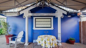 A covered patio with a blue exterior, a table with a lemon tablecloth, two white chairs, and potted plants. - Gratitude Inn - Pacific Grove House - Downtown - Close to Beach (Pacific Grove)