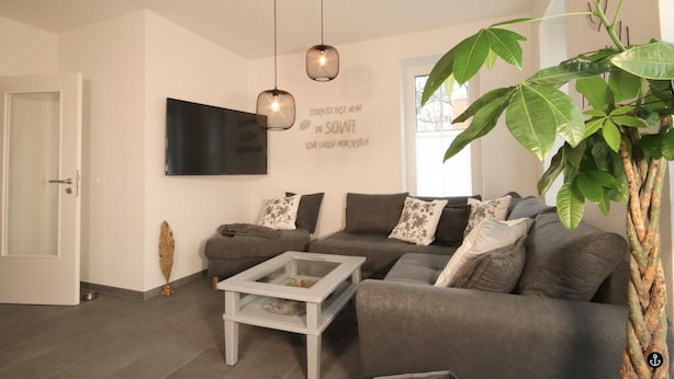 A modern living room with a sectional sofa, a glass coffee table, and a large plant. - Ferienhaus am Angerplatz (Cuxhaven)