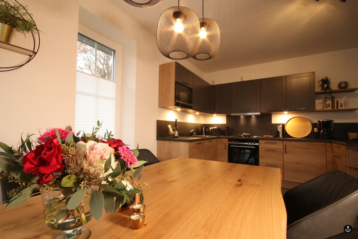 A modern kitchen with wooden cabinets, a dining table with a vase of flowers, and pendant lighting.