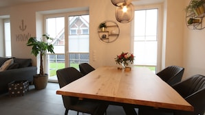 A dining area with a wooden table, black chairs, a potted plant, and a wall-mounted shelf with decorative items. - Ferienhaus am Angerplatz (Cuxhaven)