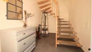 A staircase with wooden steps and a glass railing, a white dresser with drawers, a mirror with a yellow ribbon, and a vase with dried plants on the wall. - Ferienhaus am Angerplatz (Cuxhaven)