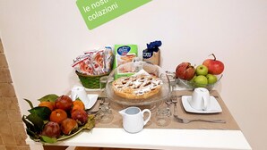 Una mesa de desayuno con un frutero, un plato de frutas frescas, una jarra de vidrio, un plato con postre y varios productos alimenticios envasados.