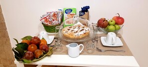 A breakfast table with a fruit bowl, a plate of fresh fruits, a glass pitcher, a plate with a dessert, and various packaged food items.