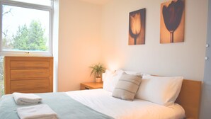 Un dormitorio con cama, una mesita de noche con lámpara, una ventana y una obra de arte en la pared.
