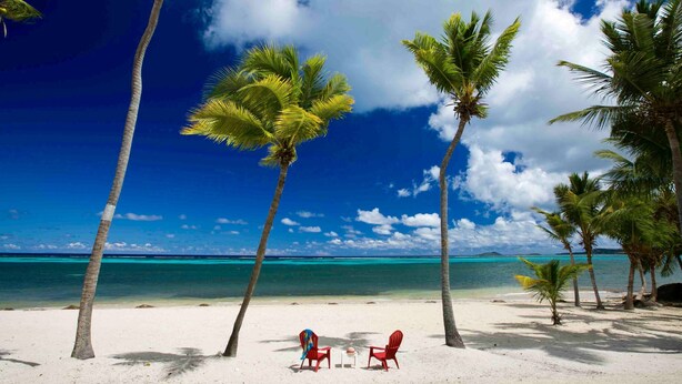 Une plage de sable blanc, bordée de palmiers, avec deux chaises rouges et une petite table.