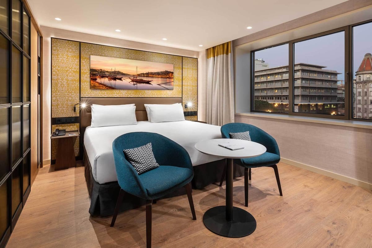 Una habitación de hotel con una cama grande, dos sillas, una mesita, un cuadro en la pared y vistas a edificios a través de la ventana.