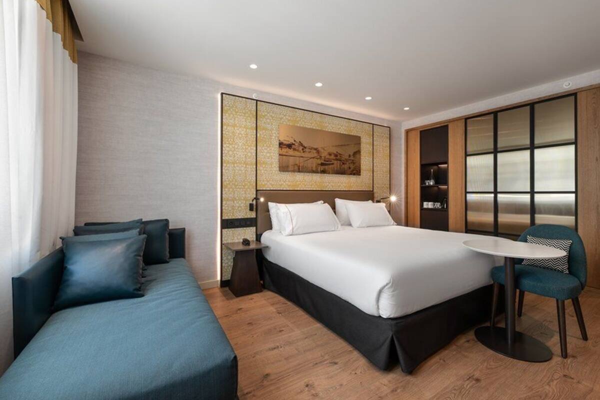 Una habitación de hotel moderna con una cama grande, un sofá, una mesita y vistas de la ciudad a través de un ventanal.