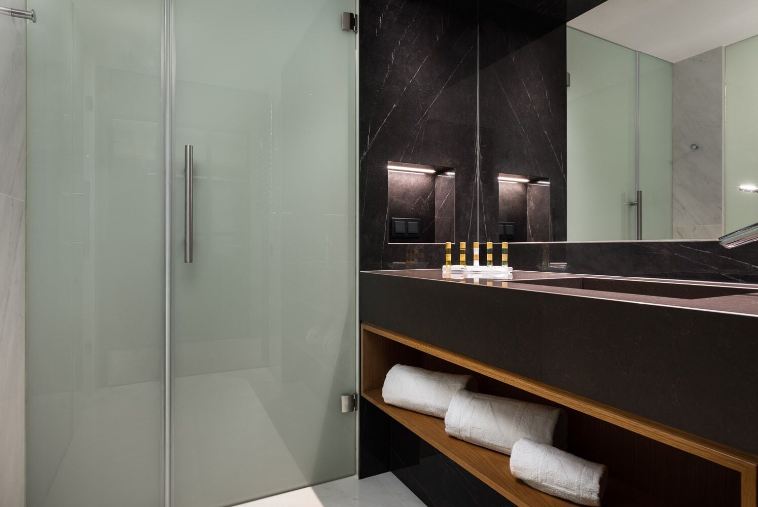 Une salle de bain moderne dotée d’une douche à parois de verre, d’un plan de toilette en marbre foncé et d’un grand miroir qui agrandit l’espace.