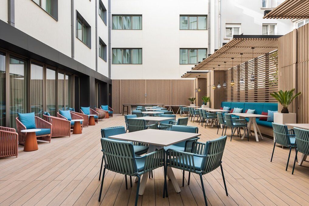 A modern outdoor seating area with tables and chairs, a wooden pergola, and a view of the building's exterior.
