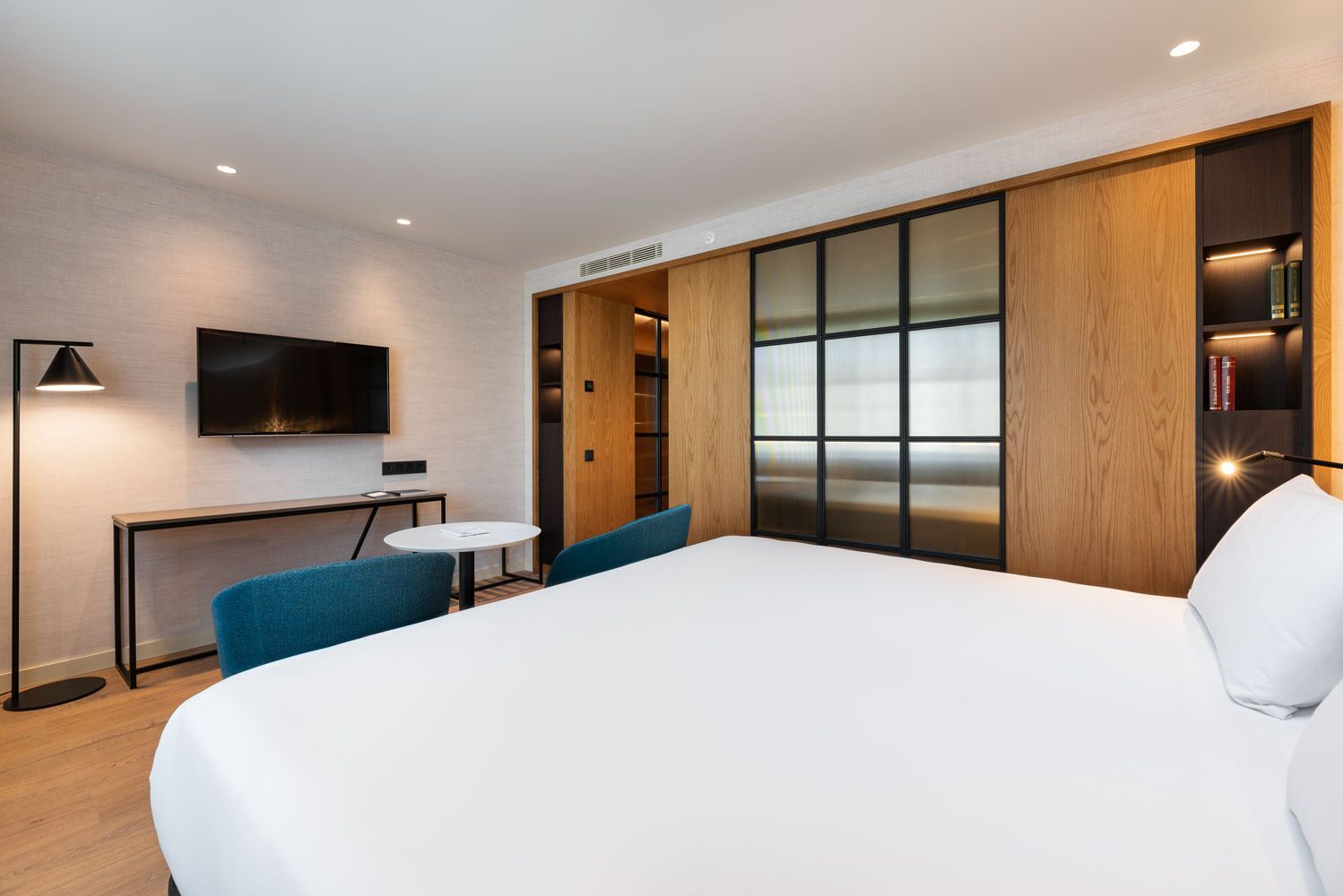 A modern hotel room with a large bed, a desk with a chair, a TV mounted on the wall, and a bookshelf.