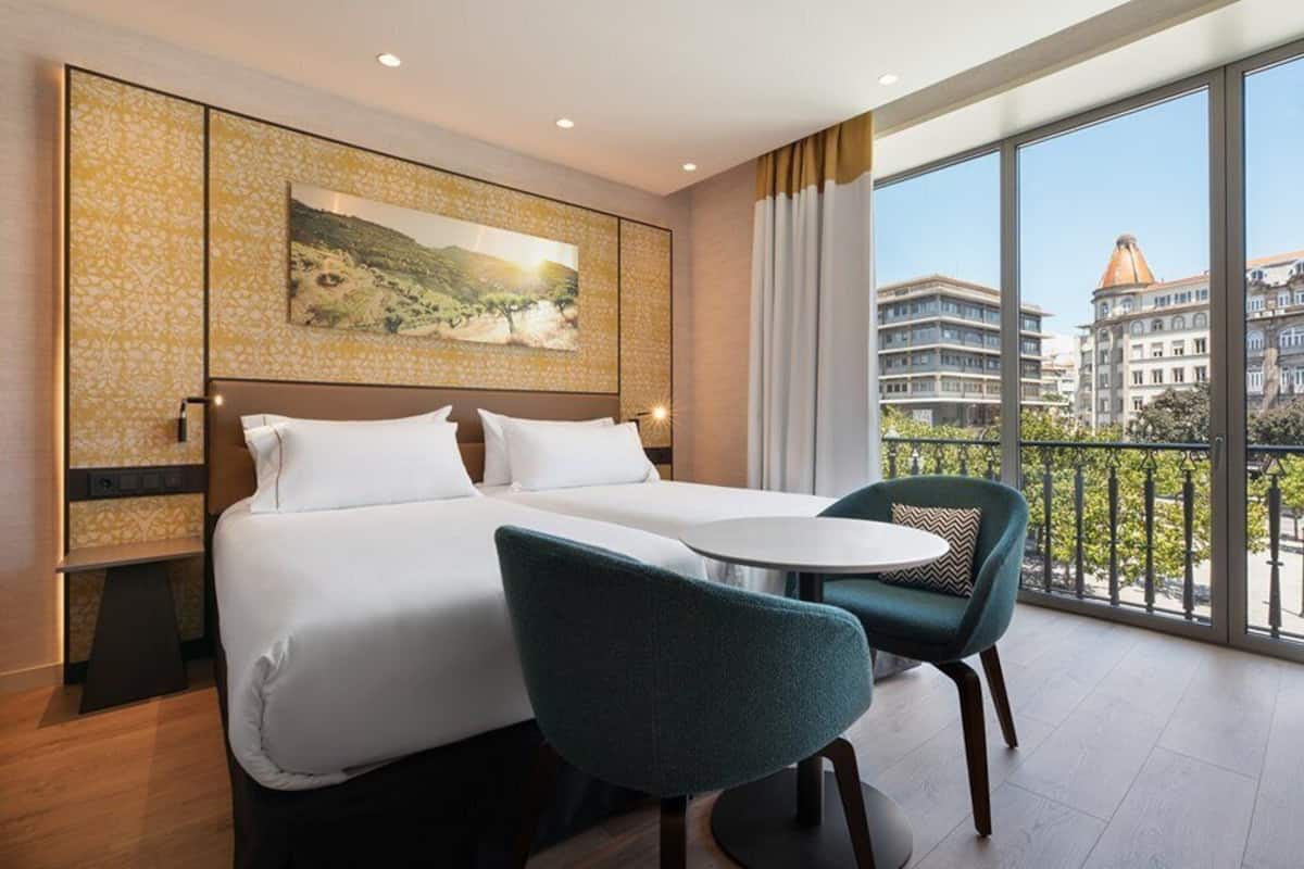 Una habitación de hotel con una cama, dos sillas, una mesita y un ventanal con vistas a la ciudad.