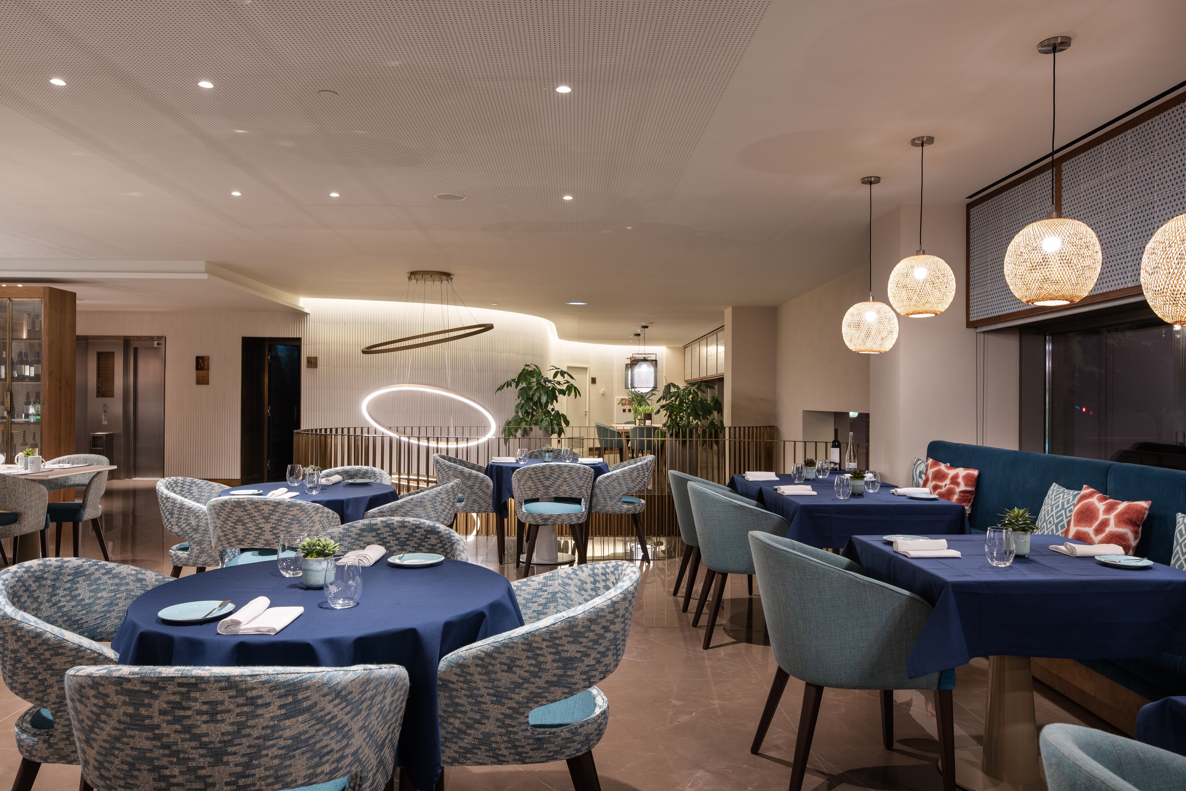Een modern restaurant met ronde tafels, blauw- en grijze gestoffeerde stoelen en hangende, sferische lampen.