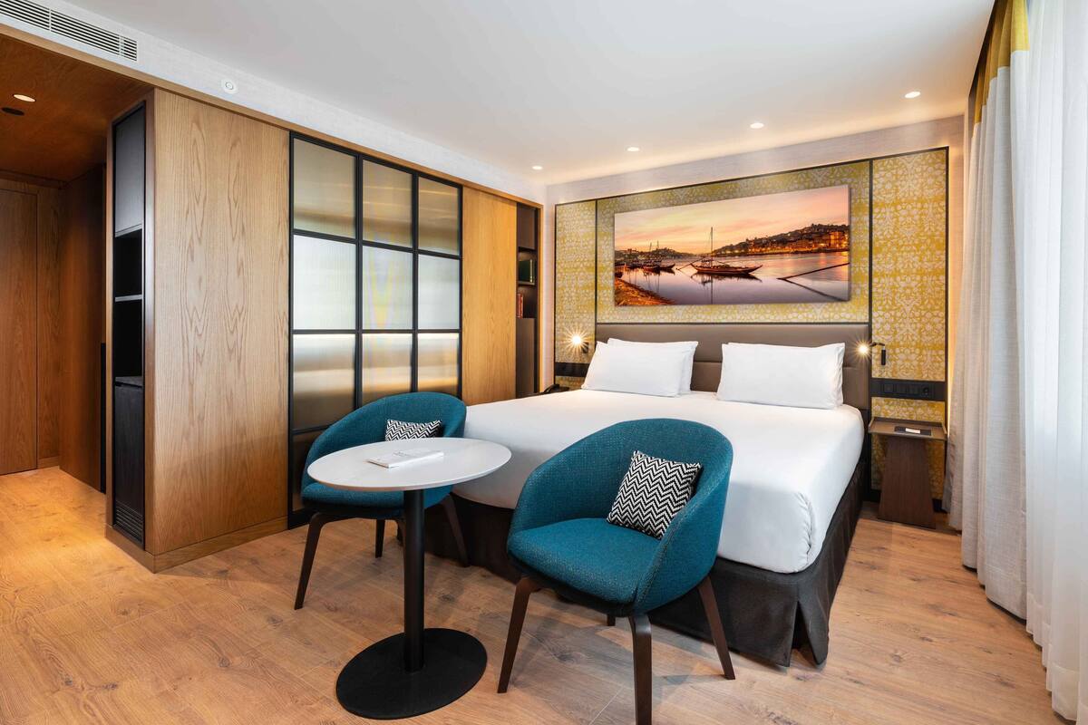 Habitación de hotel con una cama grande, dos sillas, una mesita y un cuadro de un paisaje urbano en la pared.