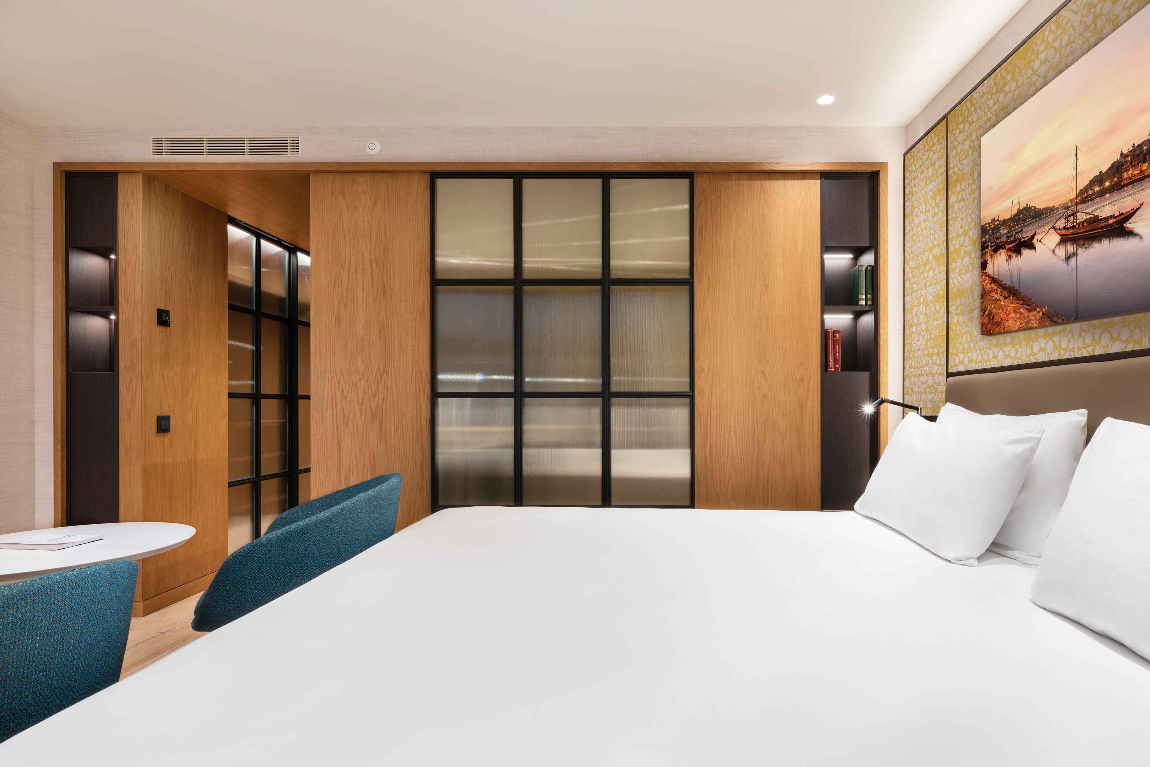A hotel room with a large bed, a seating area with a table, a bookshelf, and a large window with a view of a sunset.