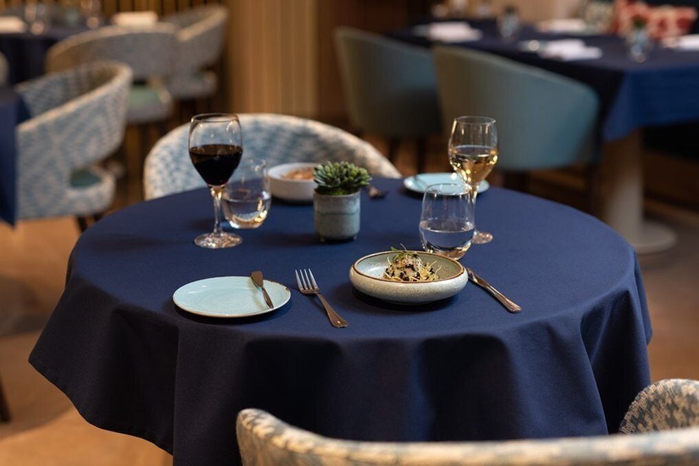 A round dining table with a dark tablecloth, two wine glasses, a plate with food, and cutlery.