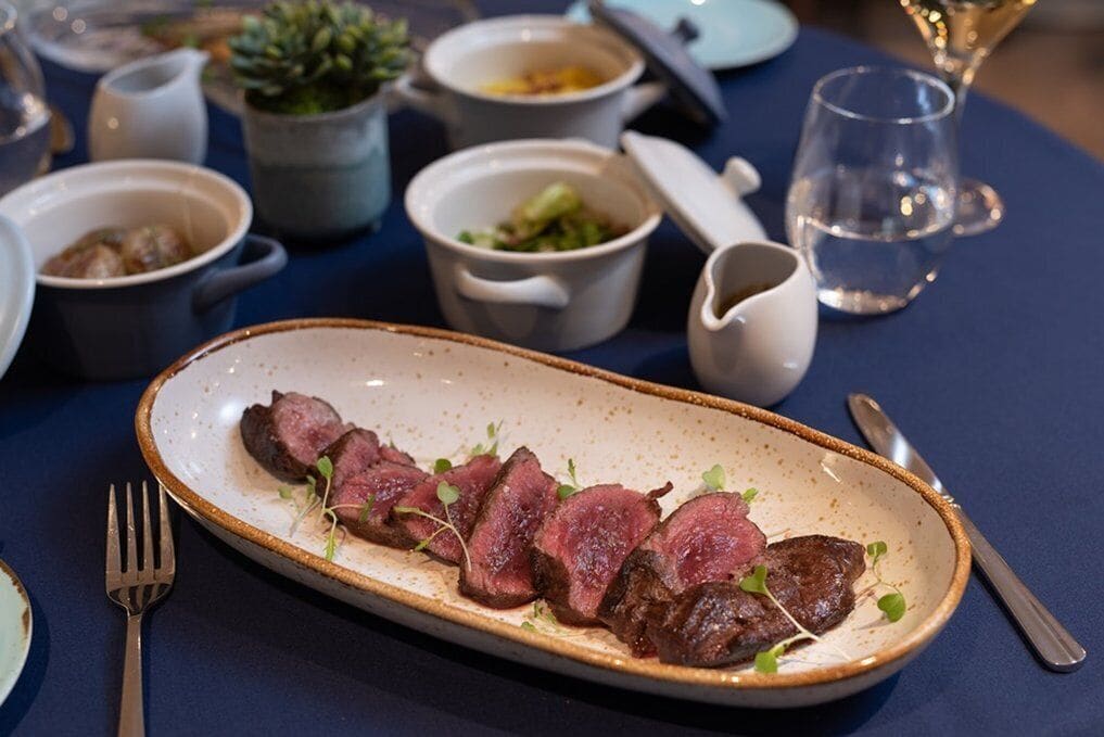 A plate of sliced steak garnished with greens, accompanied by a fork and knife on a table with various dishes and a glass of water.