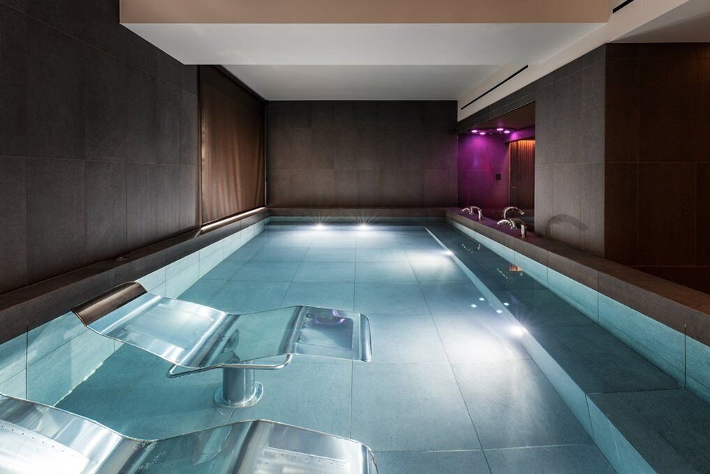 A modern indoor swimming pool with clear glass panels, a long bench, and a purple-lit wall.
