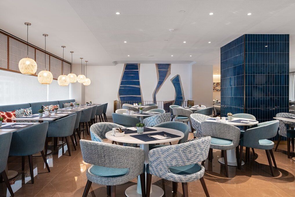 Un restaurante moderno con mesas redondas y sillas estampadas, un muro azul como detalle y lámparas colgantes.