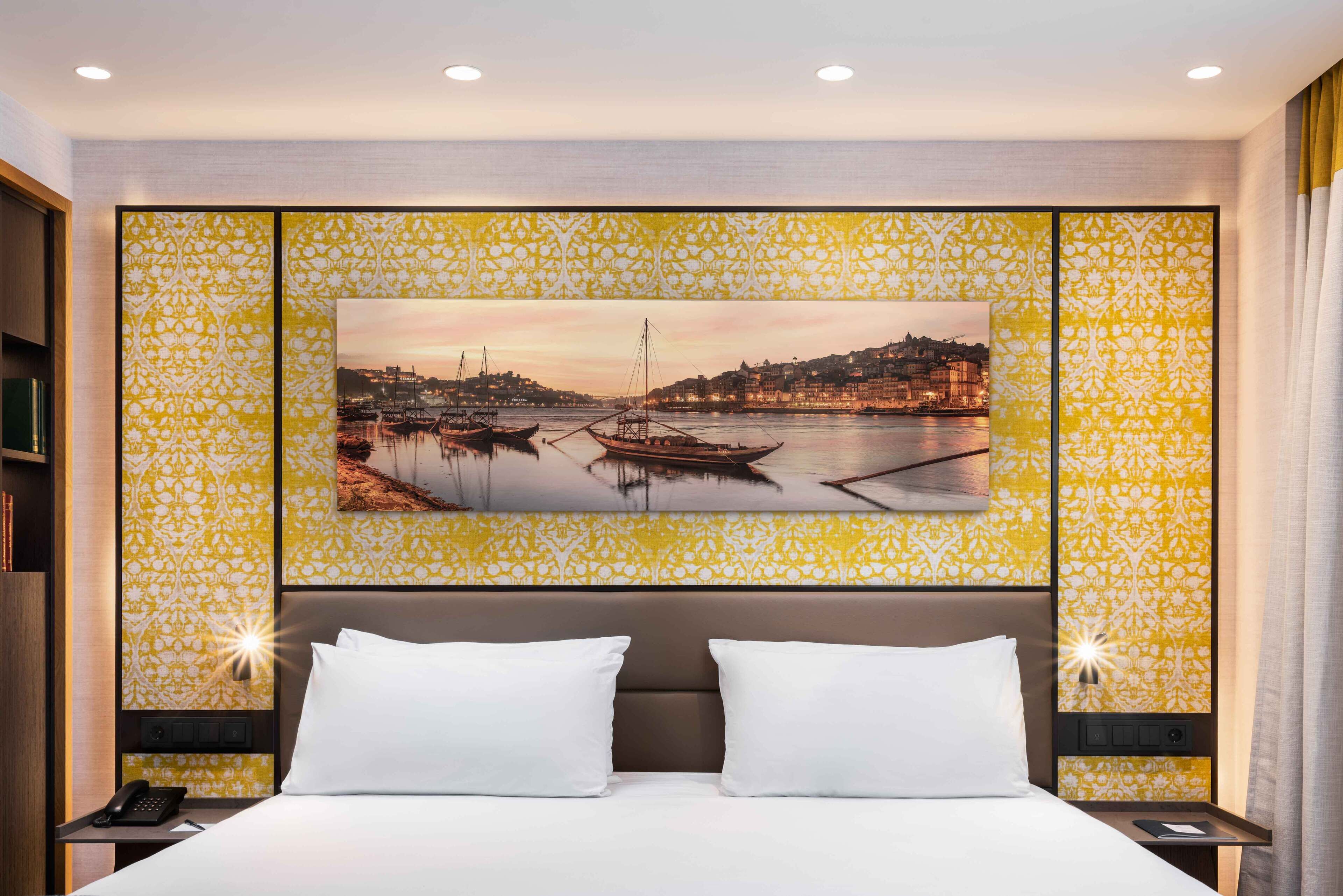 A hotel room with a bed, two white pillows, a nightstand with a phone, and a wall-mounted painting of a harbor scene.