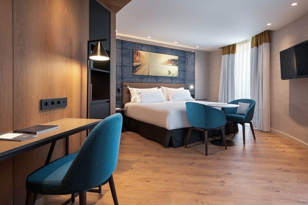 Habitación de hotel moderna con una cama, dos sillas azules, un escritorio y un televisor.