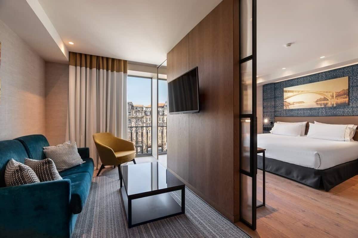 Una habitación de hotel con cama, televisor montado en la pared, sofá, sillón y vistas a la ciudad a través de amplios ventanales.