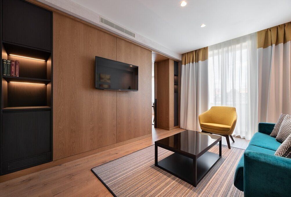 A modern living room with a wooden wall, a flat-screen TV, a coffee table, a yellow armchair, and a teal sofa.
