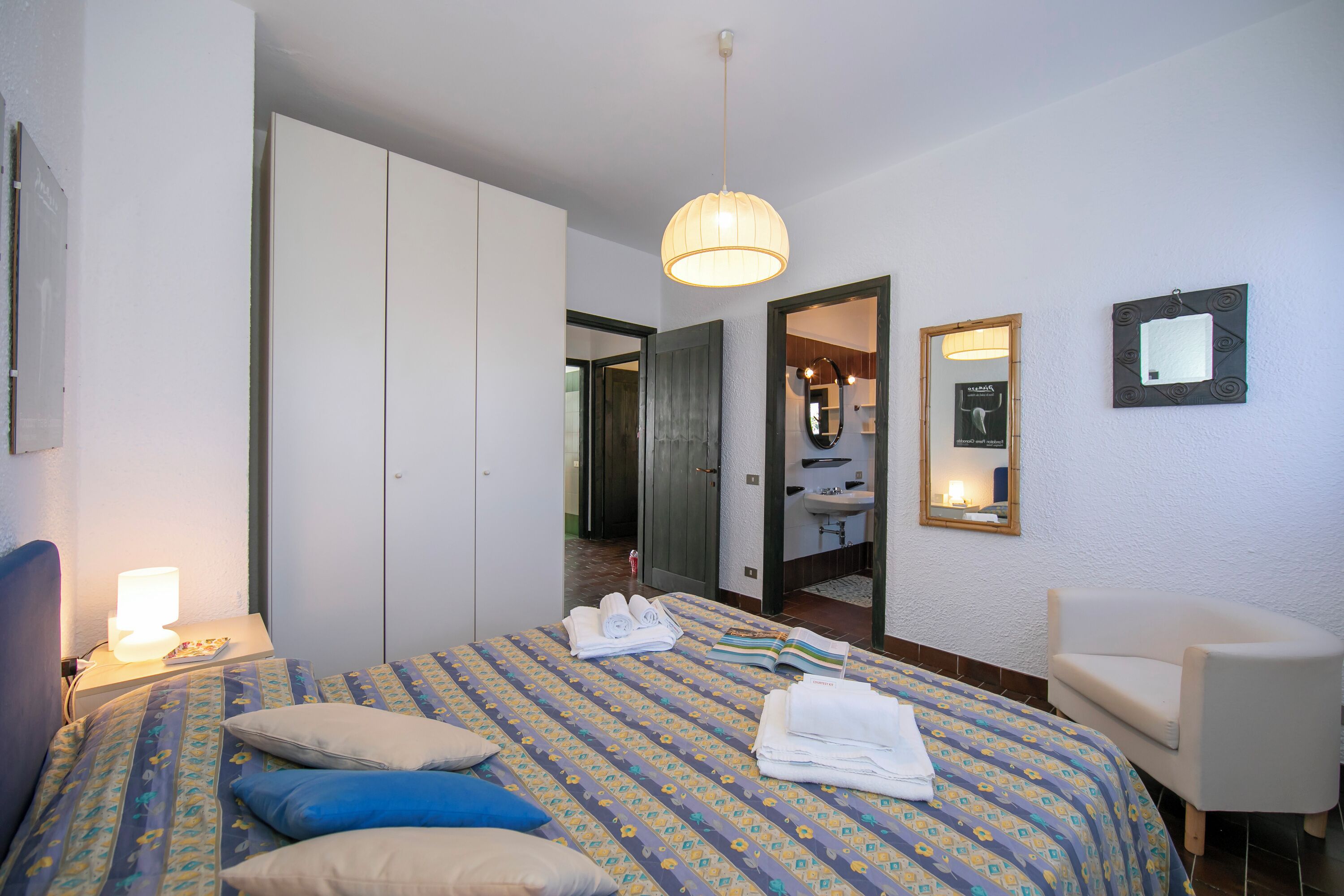 A hotel room with a bed, a chair, a wardrobe, a mirror, and a hanging light fixture.