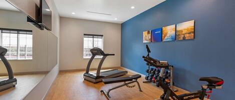 A gym with a treadmill, elliptical, and stationary bike, wooden flooring, and a blue wall with artwork.