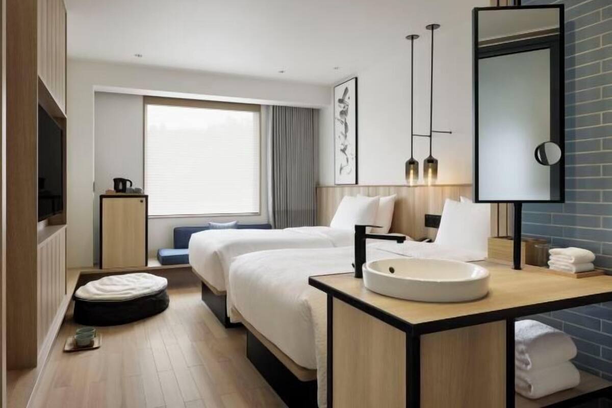 A hotel room with two beds, a large window, a round mirror, a bedside table with a sink, and a television.